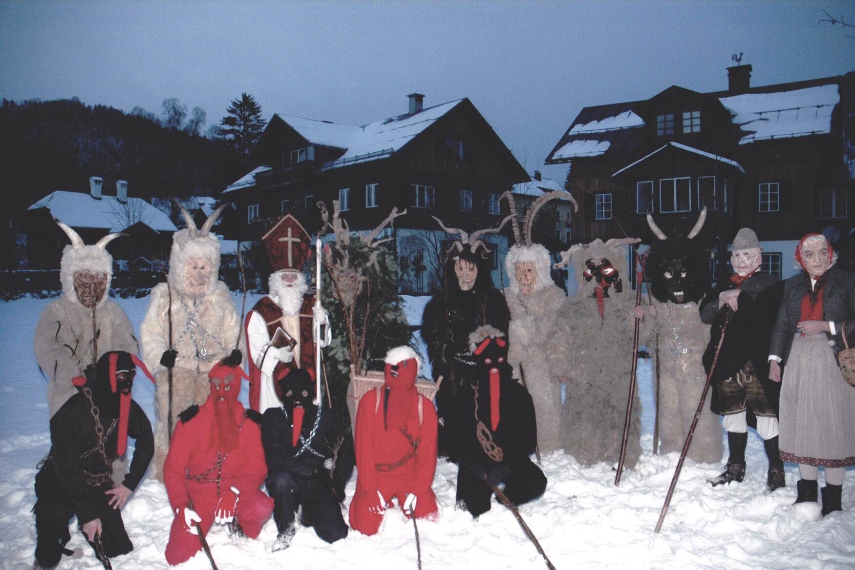 Die Miglon, wie die Krampusse im Ausseerland heißen, mit Nikolaus und Engeln vor verschneiten Häusern in Altaussee