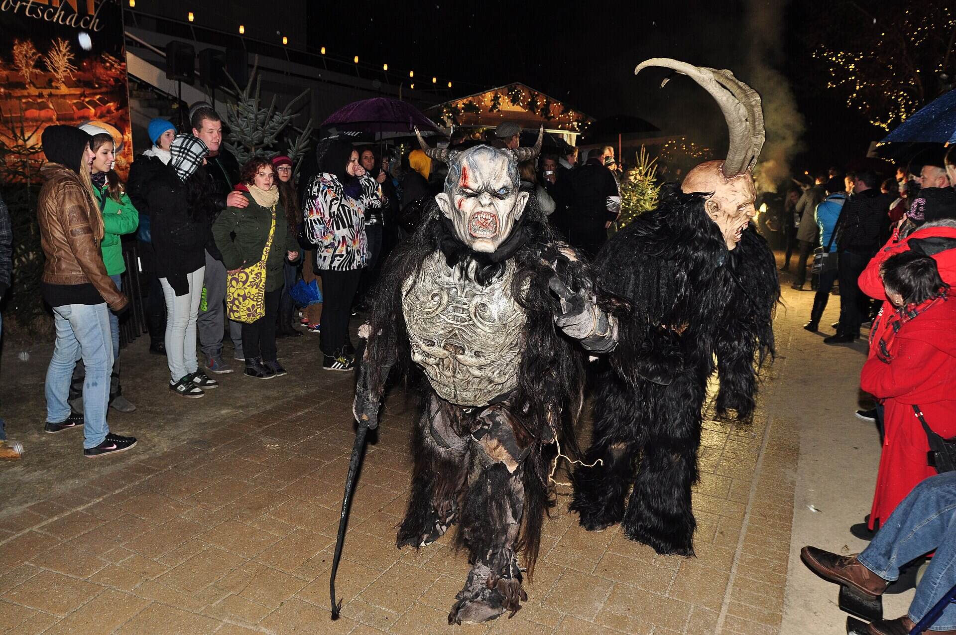Krampusse mit Hörnern, Fell und Ruten ziehen bei einem abendlichen Krampuslauf an Zuschauern vorbei